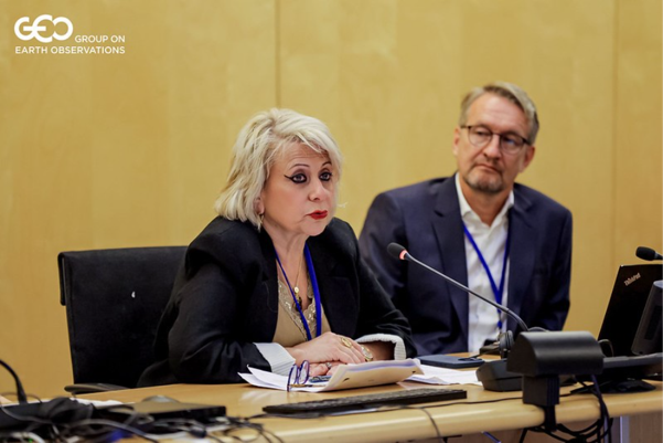 Joanna Drake.png Two people sit at a conference table with microphones and laptops; one woman is speaking while the man listens. A