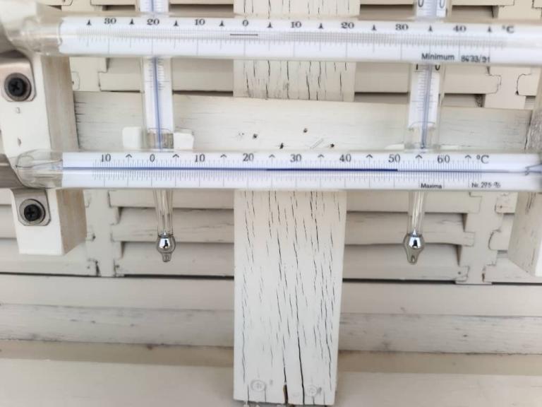Photo1.jpeg Two glass thermometers mounted on a white wooden surface, with temperature readings visible in Celsius and Fahrenheit. The wood shows visible cracks and peeling paint.