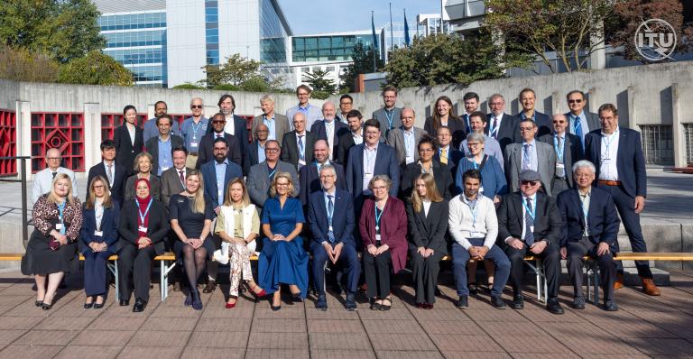 A large group of professionally dressed people poses for a formal photo outside a modern office building on a sunny day.