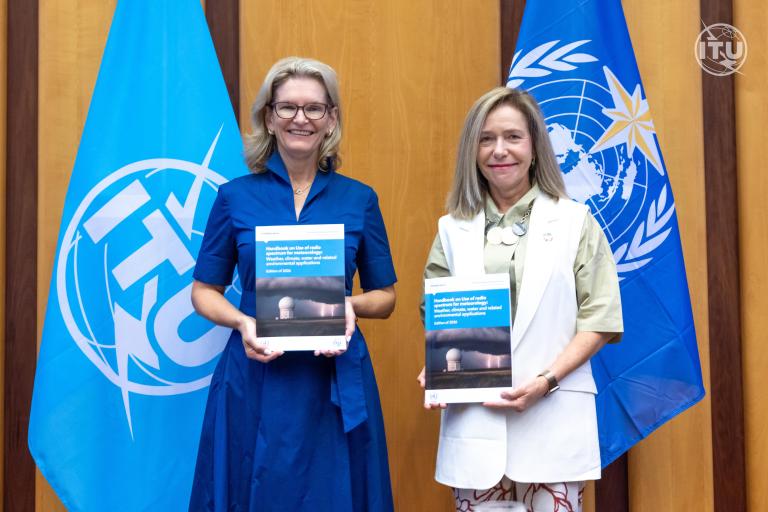 Two women stand in front of blue ITU and UN flags, holding copies of the same report and smiling at the camera.