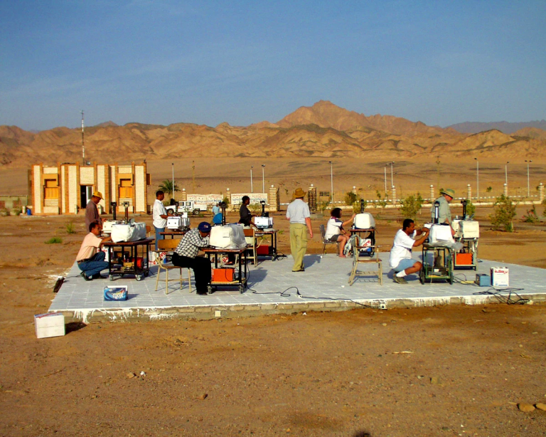 People using computers outdoors on a large mat in a desert area, with mountains and a few buildings visible in the background under a clear sky.