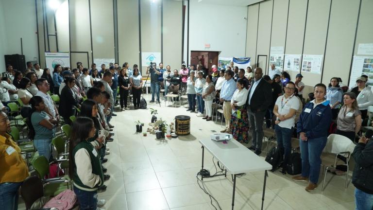 Photo 1B_Closure with Riural Indeigenous Communities in Popayan.jpg A large group of people stands in a conference room, facing each other in a circle around a display of plants and objects at the center. Posters are visible on the walls.