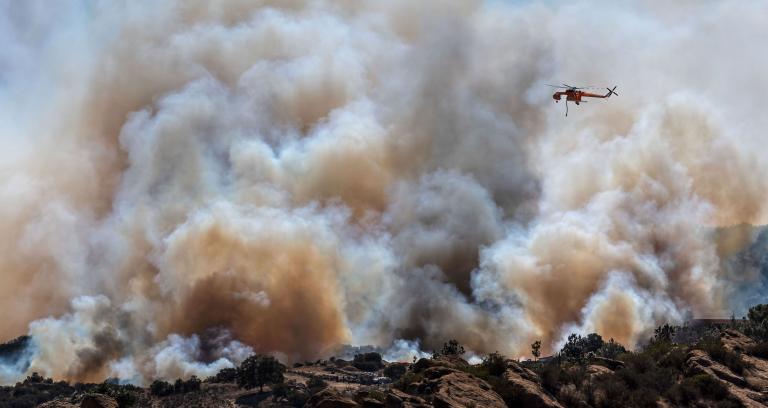 A helicopter flies over a large wildfire, releasing thick smoke into the sky above a landscape of rocks and sparse vegetation.