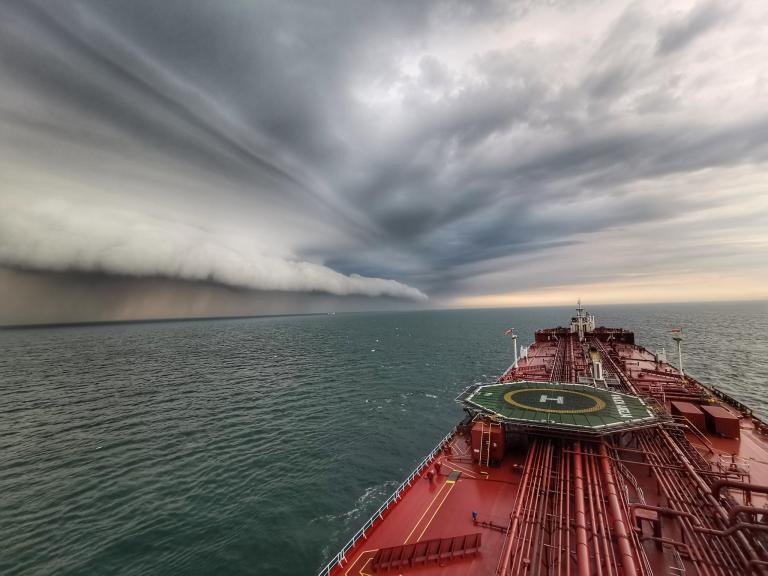 A ship sails on the ocean toward a distant rolling cloud beneath a dramatic, stormy sky.