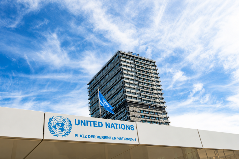 UNU.png United Nations building with logo and flag under a blue sky with clouds; address