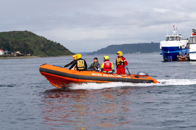 A bright orange rescue boat carrying five crew members in life vests and helmets travels across the water, with a larger vessel visible in the background.