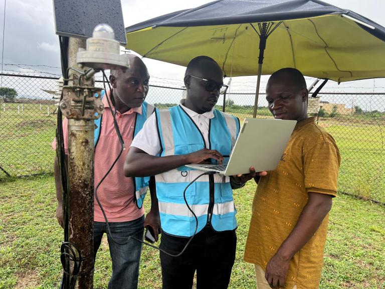 06_Liberia.jpeg Three men stand outdoors beside meteorological equipment; one in a blue vest uses a laptop under a large umbrella, while the other two observe. A chain-link fence is in the background.