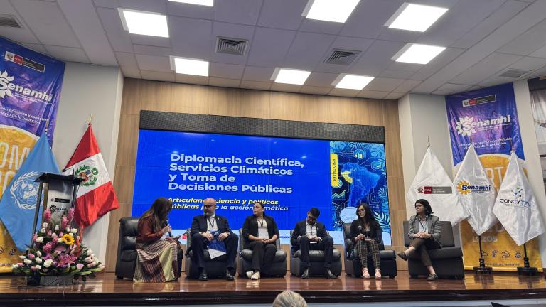 Six people sit in a row on a stage during a panel discussion, with a large screen behind them displaying the event title “Diplomacia Científica, Servicios Climáticos y Toma de Decisiones Públicas.”