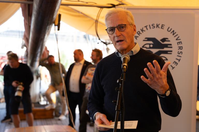 MeteoWorld 09 - Polar Year - Image 1.jpg A man with grey hair and glasses speaks into a microphone while holding a paper, with the UiT The Arctic University of Norway logo visible on a sign behind him.