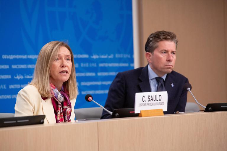 A woman and a man sit at a conference table with microphones; a nameplate reads “C. SAULO” in front of the man.