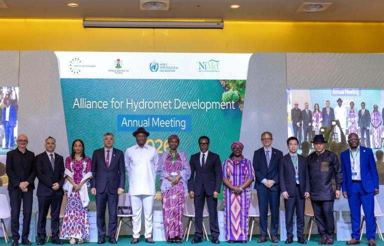 2026 Annual Meeting in Lagos, Nigeria Thirteen people stand in a row on stage in front of a projection screen for the Alliance for Hydromet Development Annual Meeting.