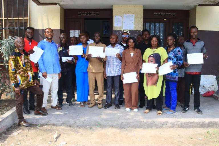 A group of adults standing in front of a building entrance, each holding a certificate.