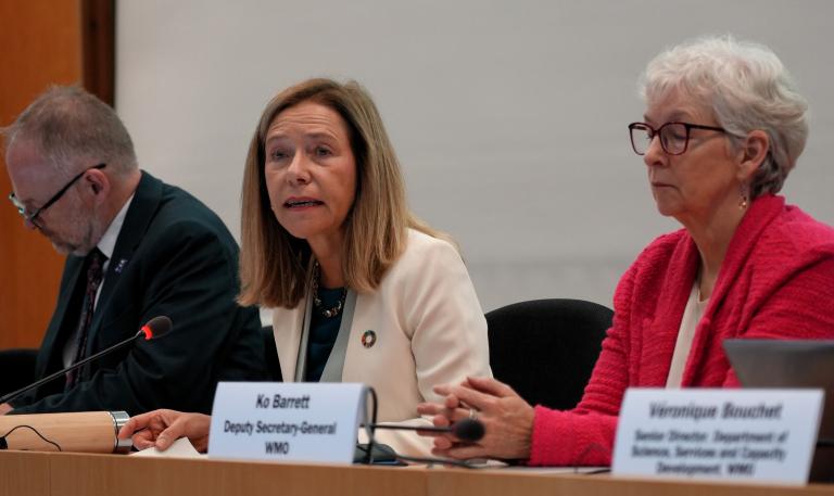 Three people seated at a table with nameplates during a meeting; Ko Barrett sits in the center.
