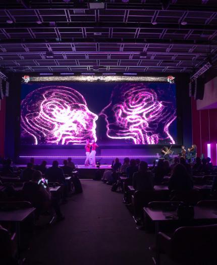 A speaker presents on a stage with a large screen displaying neon graphics of two human heads in profile. The audience is seated and attentive in a dimly lit auditorium.