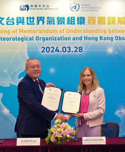 Two individuals exchanging a signed memorandum at a ceremony with a backdrop displaying logos of the world meteorological organization and hong kong observatory.