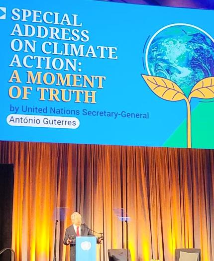 A person stands at a podium on a stage beneath a large screen displaying the text: "Special Address on Climate Action: A Moment of Truth by United Nations Secretary-General António Guterres.