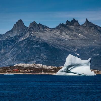 A large iceberg floats in a calm blue sea with rocky mountains in the background under a clear sky.
