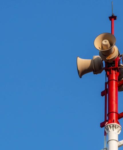 Four loudspeakers mounted on a red and white pole against a clear blue sky.