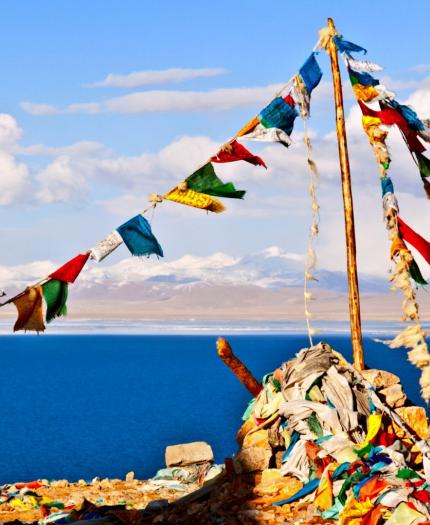 Colorful prayer flags strung between poles on a rocky hill overlooking a deep blue lake with snow-capped mountains in the distance under a partly cloudy sky.