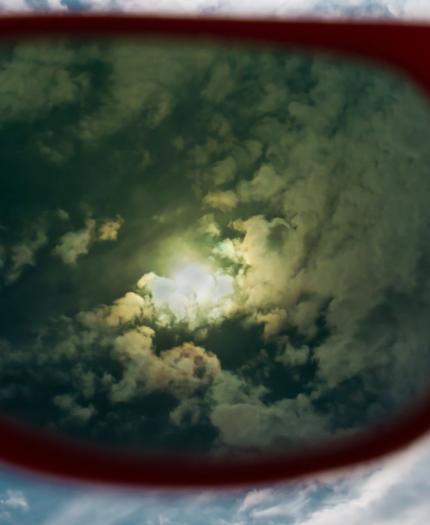 View of cloudy sky with a bright sun seen through red-framed sunglasses.