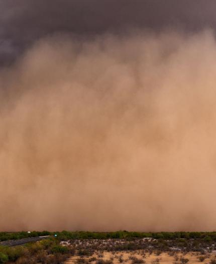 A large dust storm sweeps across a barren, desert landscape with a road on the left side of the image. The sky is dark and ominous.