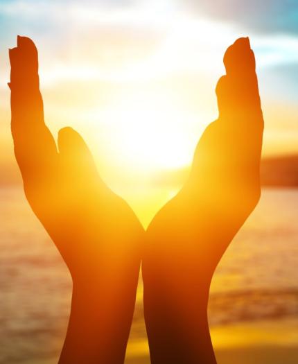 Hands are cupped together in front of a sunset over a beach, framing the sun. The sky has vibrant shades of orange, yellow, and blue.