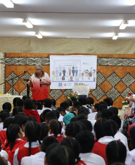 A speaker addresses a group of schoolchildren seated on the floor in a decorated room with a projected presentation behind them. Several adults are standing, and a cameraman is filming the event.