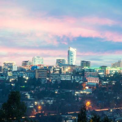 A brightly lit cityscape at dusk with a prominent modern building in the center, surrounded by smaller buildings under a sky with pink and blue hues.