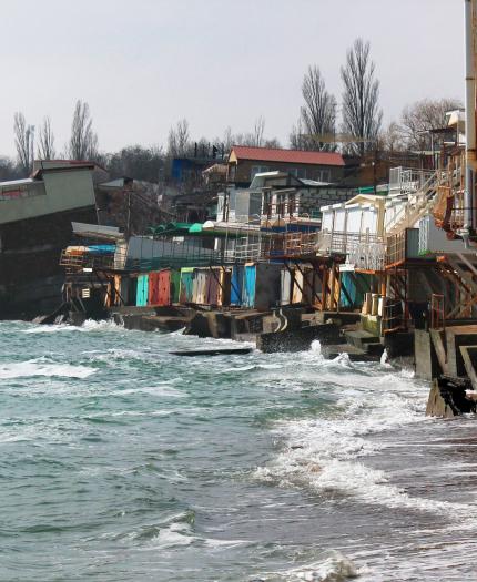 Beachfront buildings extensively damaged by erosion with portions collapsed into the sea, and waves lapping against the remains of the structures. Trees are visible in the background.