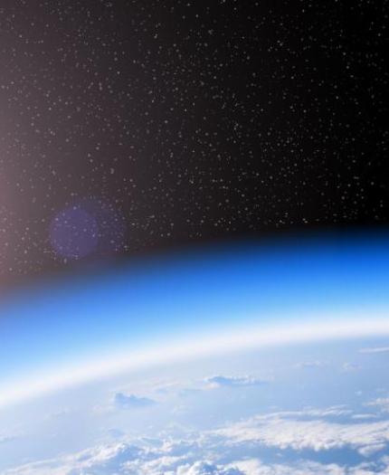 Image of Earth’s curved horizon as seen from space with the sun shining in the upper left and stars visible in the dark sky above. The planet's atmosphere and some cloud formations are also visible.