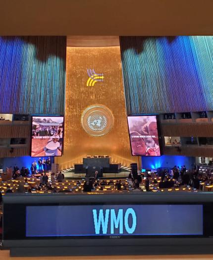 A large conference hall filled with attendees, featuring a stage with a large emblem and multiple screens displaying images. The foreground shows a sign with "WMO.