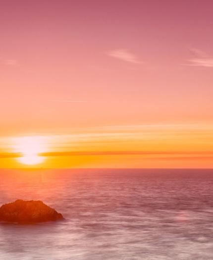 A serene sunset over a calm ocean with two small rocks in the foreground, under a pink and orange sky.