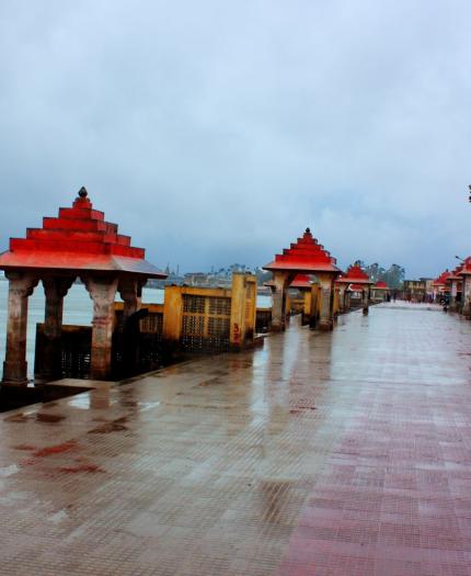 A riverside promenade with red-roofed pavilions on a rainy day. The ground is wet, and the sky is overcast. The walkway is empty except for a lone dog.