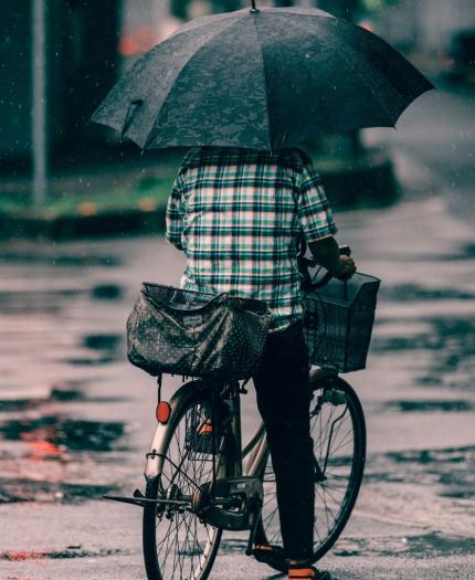 Person riding a bicycle in the rain, holding a large black umbrella and carrying two bags on the bike. The street is wet with visible puddles.