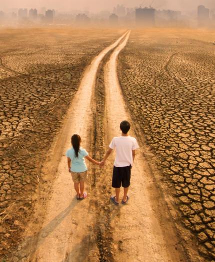 Two people stand on a dry, cracked path leading toward a distant city skyline under a hazy sky.