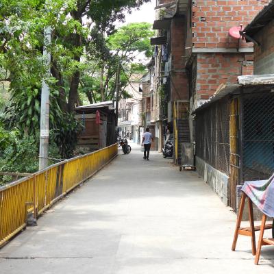 Narrow alley with brick buildings on one side, trees on the other, a yellow railing, and a person walking away in the distance.
