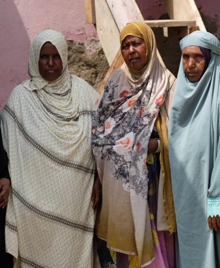 Five women stand side by side, wearing various colored hijabs. The background features a pink wall and a staircase.