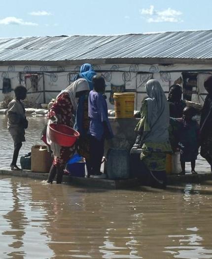 People gather with containers on a raft in floodwater near a building with a metal roof.