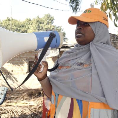 A woman in a gray headscarf and orange cap speaks into a megaphone outdoors, near a tree and some debris.