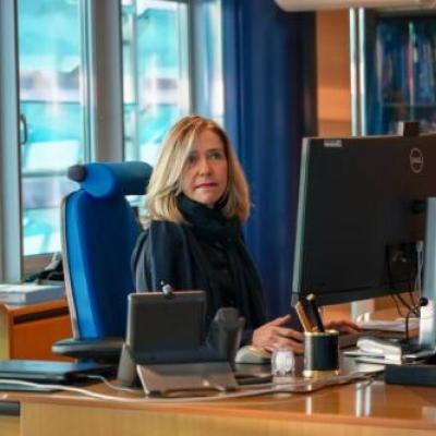 A person sitting at a desk, working on a computer in an office. A United Nations flag and bookshelf are visible in the background.