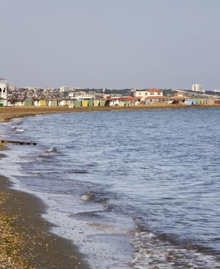 A sandy beach with gentle waves, bordered by a town with buildings and colorful beach huts in the background under a clear sky.