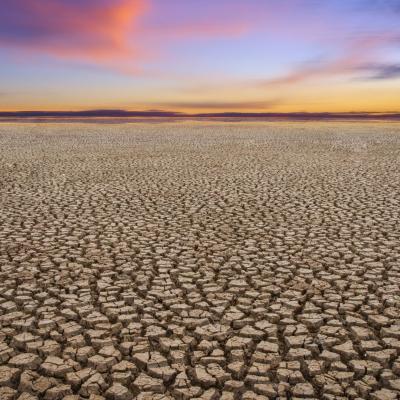 Cracked dry earth stretches toward a colorful sunset sky with pink and orange clouds on the horizon.