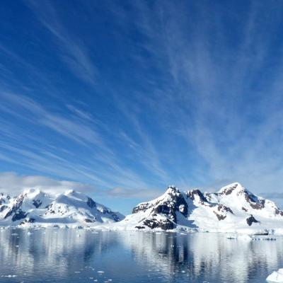 Snow-covered mountains and glaciers are reflected in calm Antarctic waters under a blue sky with wispy clouds.