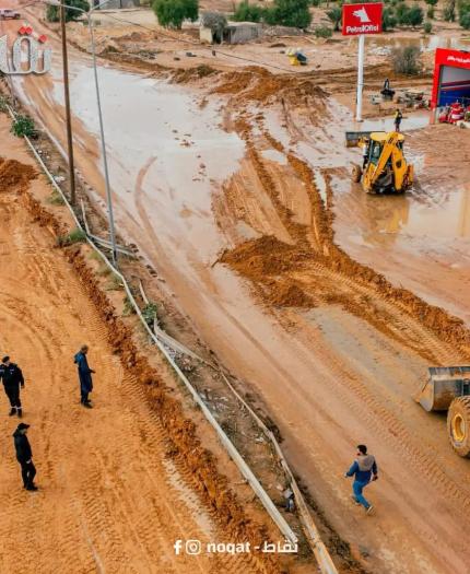 Flooded street with muddy water, people walking, and construction vehicles working near a PetroOil station.