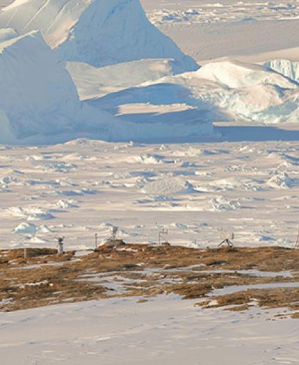 Two small structures on a barren landscape with snow and large ice formations in the background.