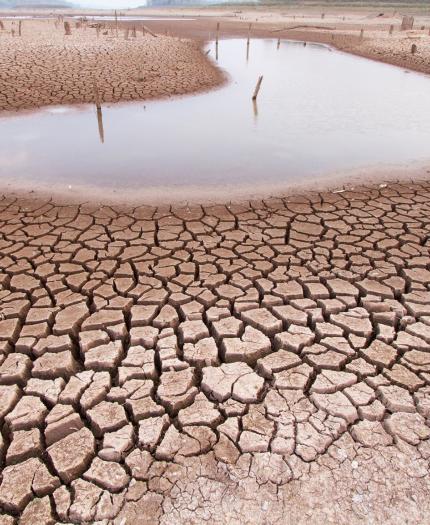 Cracked, dry earth surrounds a small pool of water in an arid landscape under a cloudy sky.