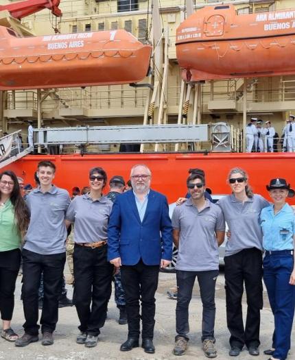 A group of people stands in front of a red ship with lifeboats. The ship's name includes "Irizar" and "Buenos Aires." Some individuals wear uniforms, and a car is visible nearby.