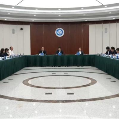A formal meeting with over twenty people seated around a large U-shaped table in a conference room, with microphones and nameplates arranged in front of them.