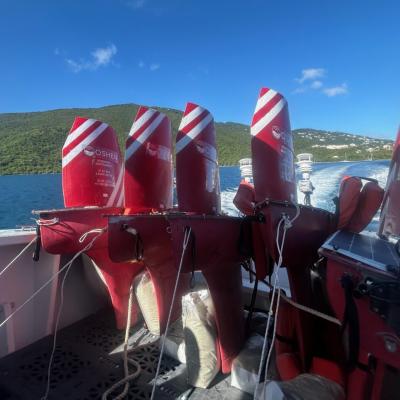 Several red and white Wave Glider marine drones are secured on the back of a boat, with the ocean and a green, hilly coastline visible in the background.