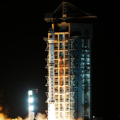 A rocket launches at night from a lit-up launch pad, emitting bright flames and smoke at the base against a dark sky.